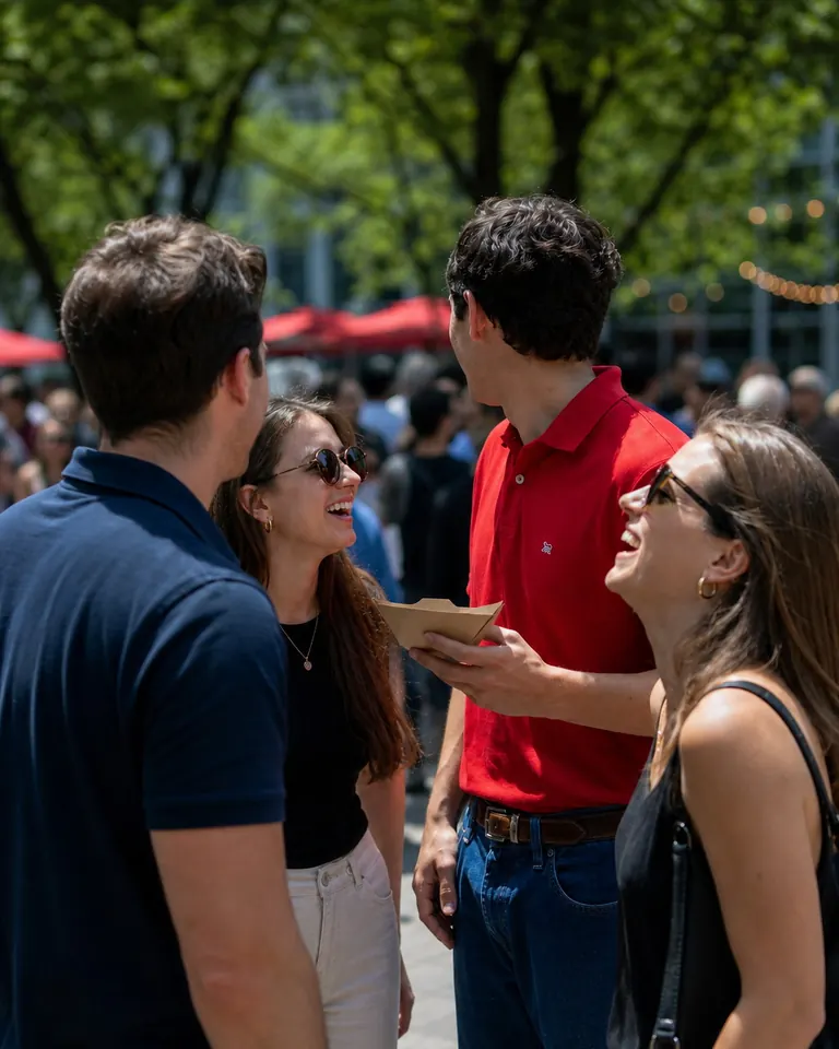 Four colleagues standing and laughing together at an outdoor lunch gathering in a tree-lined Downtown DC plaza.